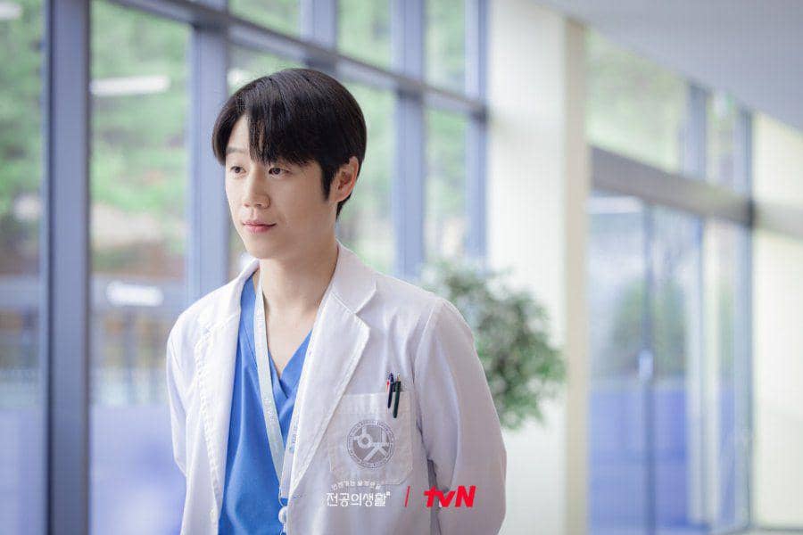 Seorang dokter muda mengenakan jas putih dan seragam medis biru berdiri di koridor rumah sakit dengan latar jendela besar.