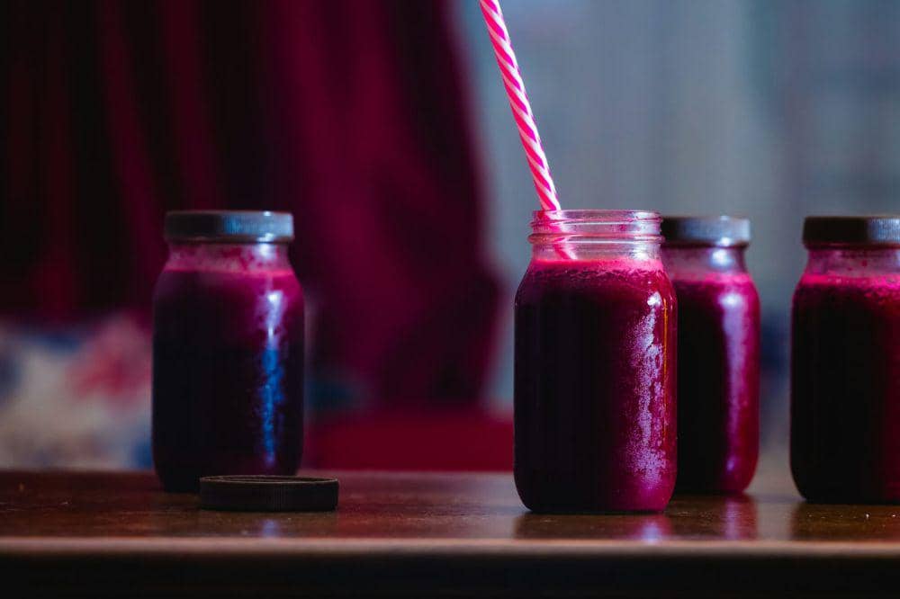 Beberapa botol berisi jus bit berwarna ungu kemerahan di atas meja kayu dengan satu botol memiliki sedotan bergaris merah putih.