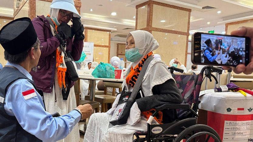 Seorang jemaah haji lanjut usia duduk di kursi roda berbincang dengan petugas haji di ruang tunggu bersama jemaah lainnya.