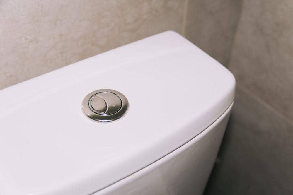 Tombol flush toilet berwarna krom di atas tangki toilet putih dengan latar dinding keramik abu-abu di kamar mandi.