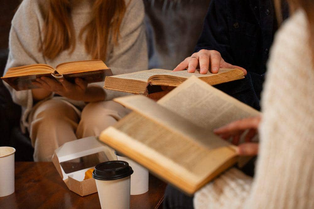 Beberapa orang duduk bersama sambil membaca buku tebal di meja kayu dengan kopi dan kotak makanan di sekitarnya.
