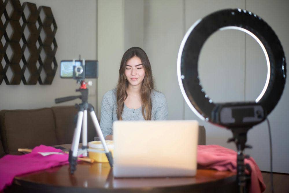 Seorang perempuan duduk di depan laptop dengan lampu ring light dan kamera ponsel di tripod saat membuat konten di rumah.