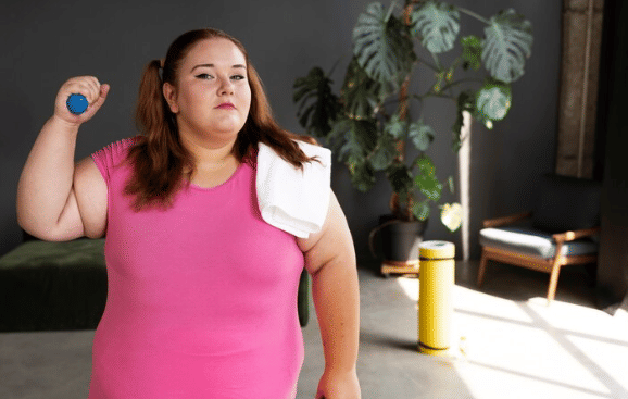 Perempuan bertubuh besar mengenakan kaus pink memegang dumbbell kecil dan handuk di bahu, berdiri percaya diri di ruang olahraga.