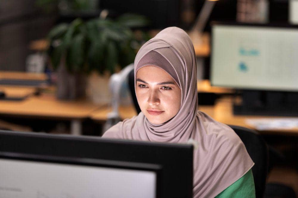 Seorang perempuan berhijab bekerja dengan fokus di depan komputer di ruang kantor modern dengan pencahayaan hangat.