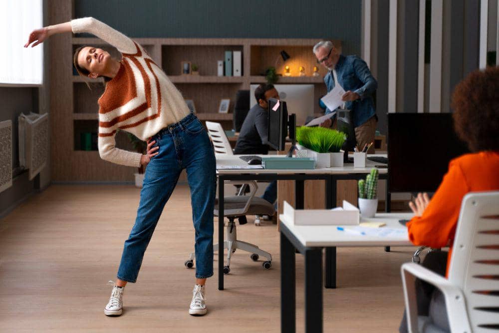 Seorang pekerja kantor melakukan peregangan tubuh di antara meja kerja sambil rekan-rekannya beraktivitas di ruang kantor modern.