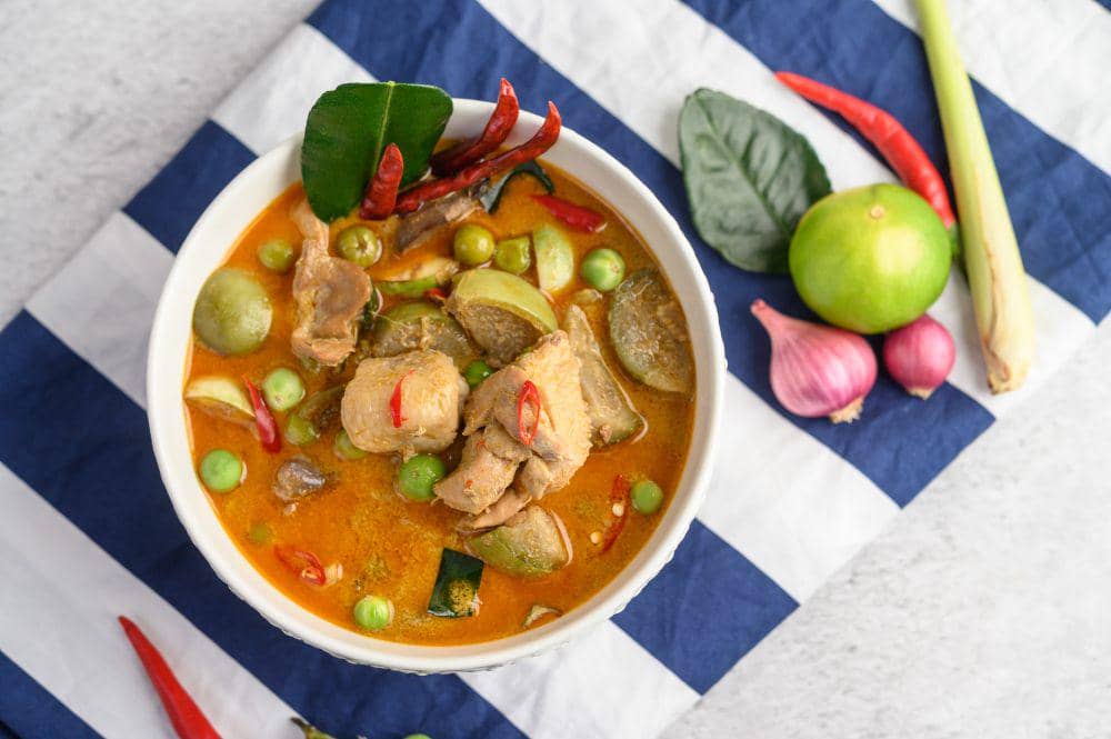 Semangkuk gulai jeroan sapi berkuah santan kuning kemerahan dengan potongan cabai, daun jeruk, dan sayuran di atas kain bergaris biru putih.