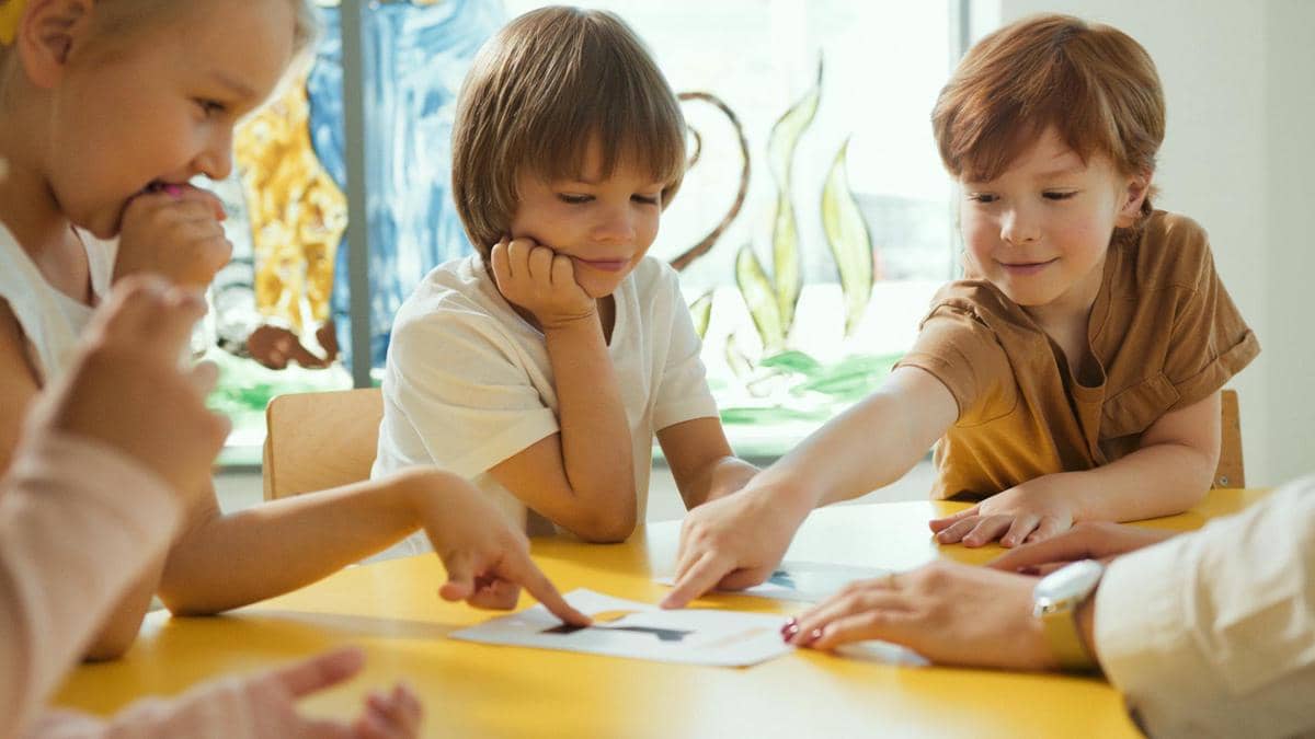 Beberapa anak duduk di meja kuning sambil berdiskusi dan menunjuk gambar di atas kertas di ruang kelas yang cerah.