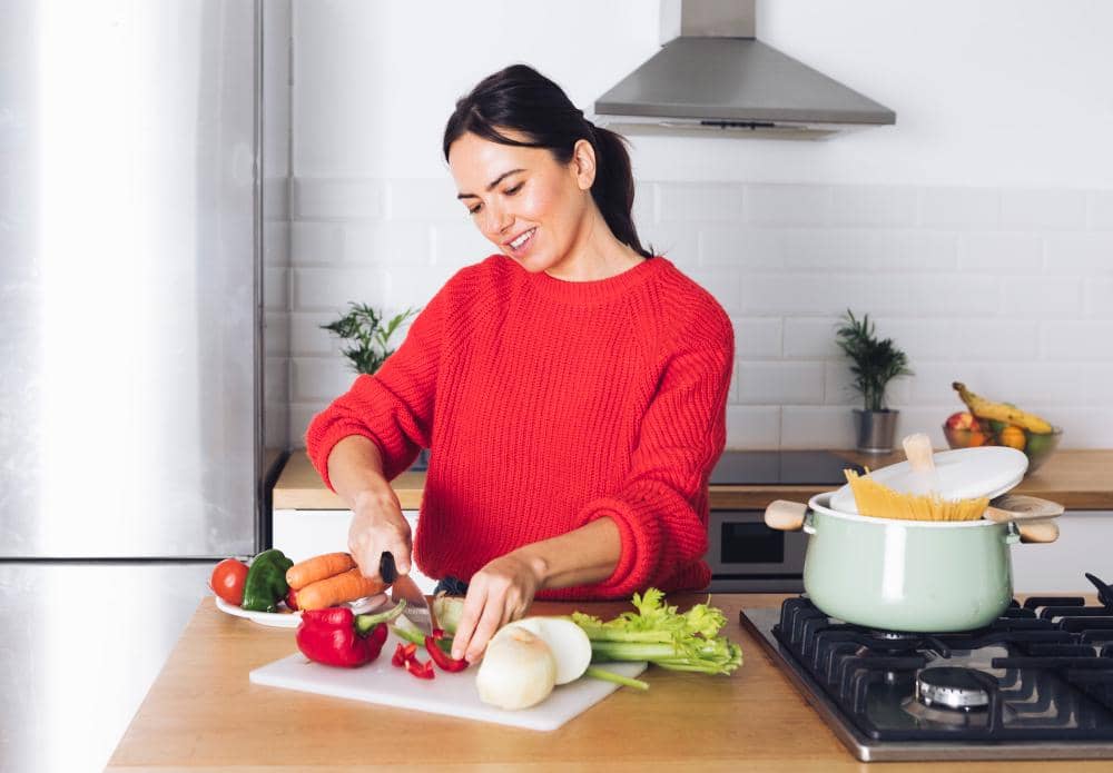 Seorang wanita mengenakan sweter merah sedang memotong sayuran di dapur modern dengan panci berisi pasta di atas kompor.