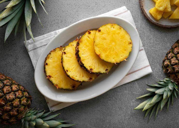 Irisan buah nanas segar berwarna kuning disajikan di atas piring oval putih dengan latar meja abu-abu dan beberapa nanas utuh di sekitarnya.