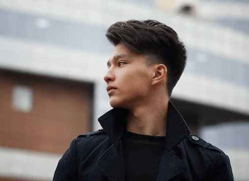 Seorang pria berambut gaya side part klasik mengenakan jaket hitam, berdiri di luar ruangan dengan latar belakang gedung modern.