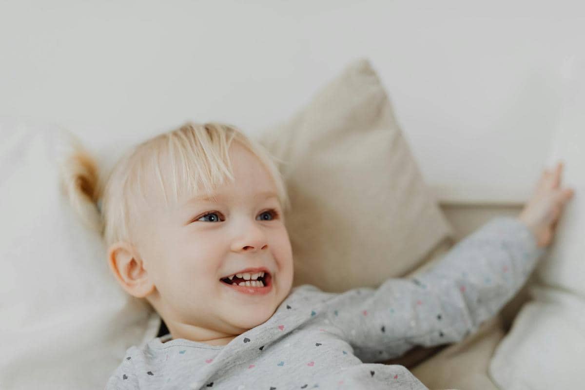 Anak kecil berambut pirang tersenyum ceria sambil berbaring di atas bantal berwarna terang dengan suasana hangat dan lembut.