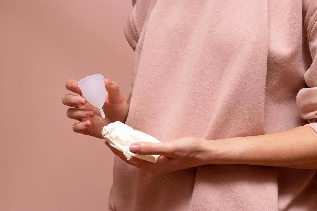 Seseorang mengenakan pakaian berwarna merah muda memegang menstrual cup berwarna putih dan kantong kain kecil di tangannya.