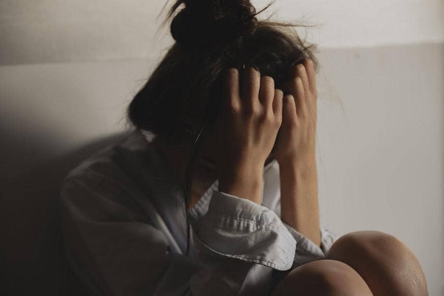 Perempuan duduk di sudut ruangan dengan kepala tertunduk dan tangan menutupi wajah, menggambarkan perasaan tertekan dan terisolasi.