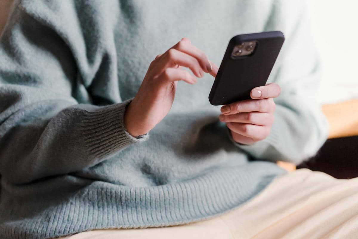Seseorang mengenakan sweter abu-abu sedang memegang ponsel dan mengetuk layar untuk mengecek saldo rekening bank.