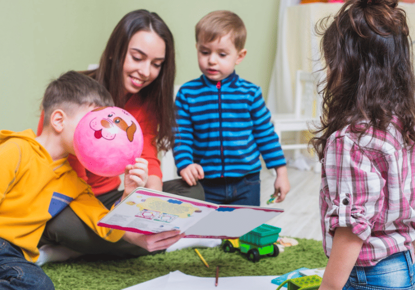 Seorang wanita muda bermain dan membaca buku bergambar bersama beberapa anak kecil di dalam ruangan yang cerah dan hangat.