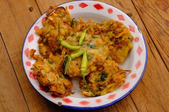 Beberapa perkedel jagung goreng berwarna keemasan disajikan di piring enamel bermotif merah putih dengan cabai hijau di atasnya.