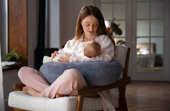Seorang ibu duduk di kursi sambil menyusui bayinya dengan bantal menyusui di pangkuannya di dalam ruangan yang hangat dan tenang.