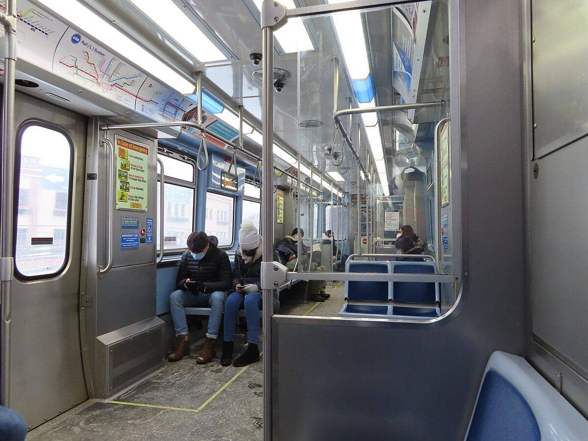 Penumpang duduk di dalam kereta L dengan interior logam dan kursi biru, beberapa orang mengenakan masker sambil menggunakan ponsel.