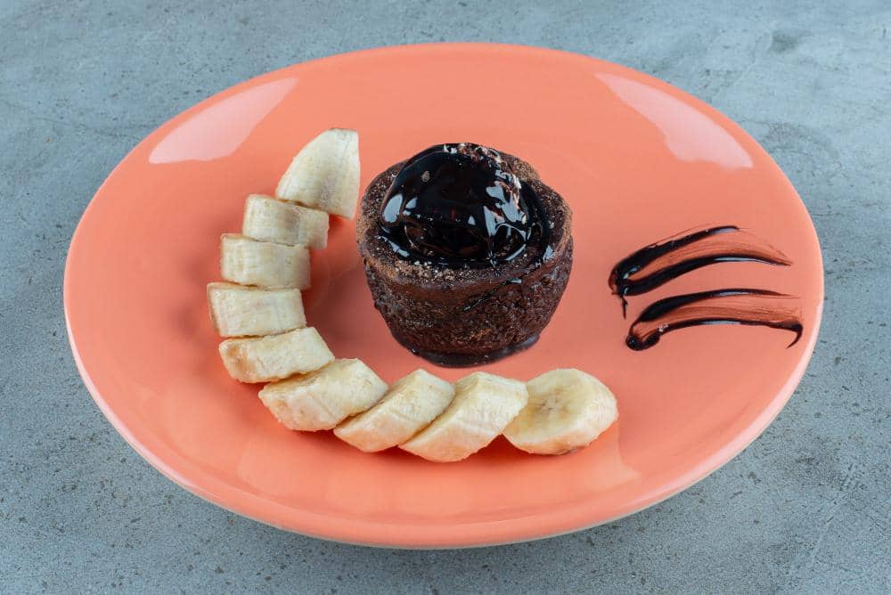 Potongan pisang tersusun melengkung di samping kue cokelat kecil dengan saus cokelat di atas piring oranye di atas meja abu-abu.