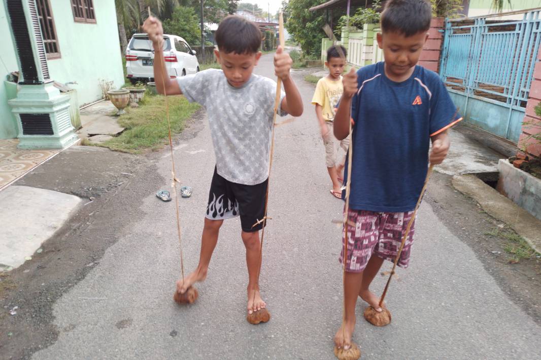 Anak-anak di sebuah perkampungan asyik bermain tradisional marsipatu hoda (terompah batok kelapa) di jalanan. Permainan sederhana ini mengajarkan keseimbangan dan kebersamaan di tengah gempuran gawai.(wikimedia.org)