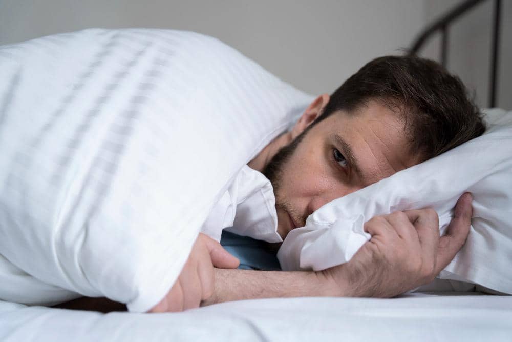 Seorang pria berbaring di tempat tidur dengan wajah terlihat lelah dan sulit tidur sambil memeluk bantal putih di kamar tidur.
