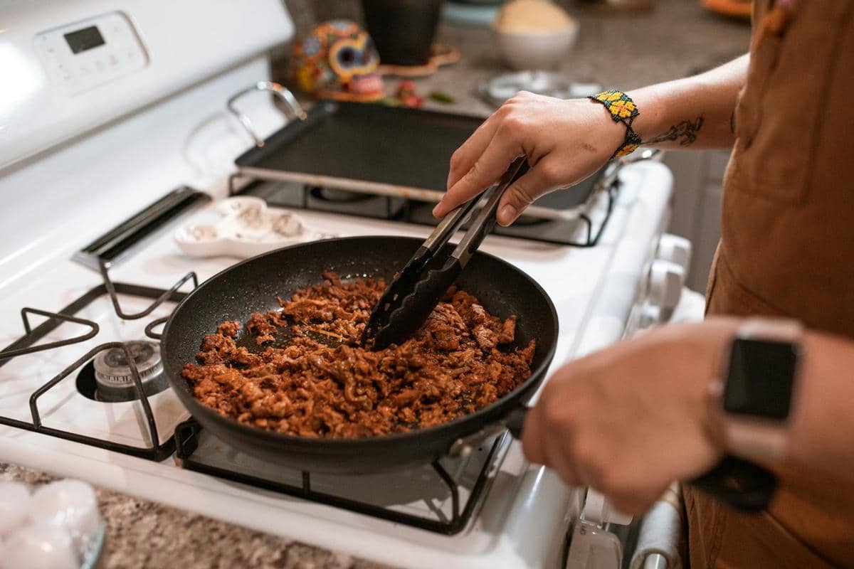 Seseorang memanaskan makanan sisa di wajan di atas kompor gas menggunakan penjepit dapur di dapur rumah.