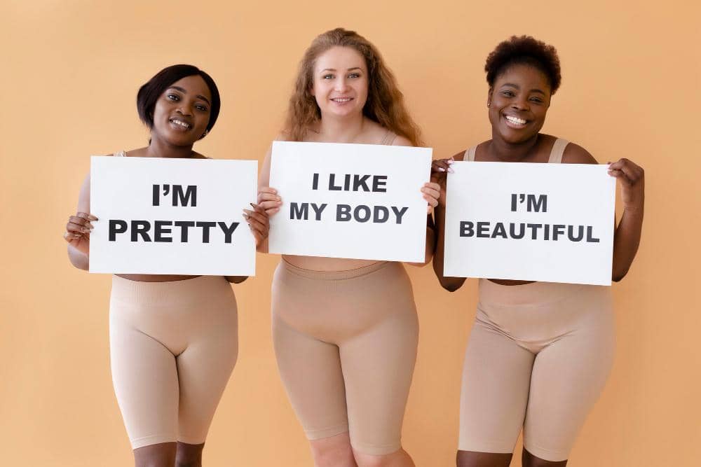 Tiga perempuan tersenyum sambil memegang papan bertuliskan pesan positif tentang penerimaan tubuh di latar oranye.