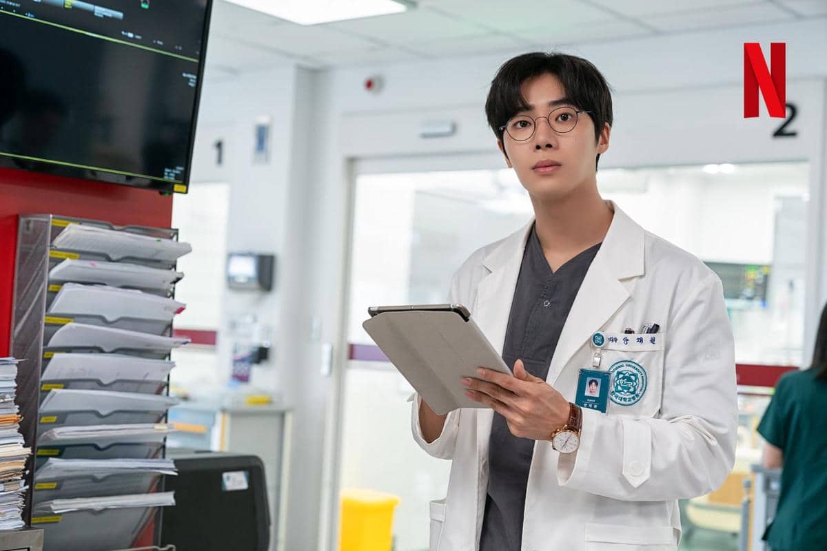 Seorang dokter muda mengenakan jas putih dan kacamata sedang memegang tablet di ruang rumah sakit dalam adegan drama The Trauma Code: Heroes on Call.