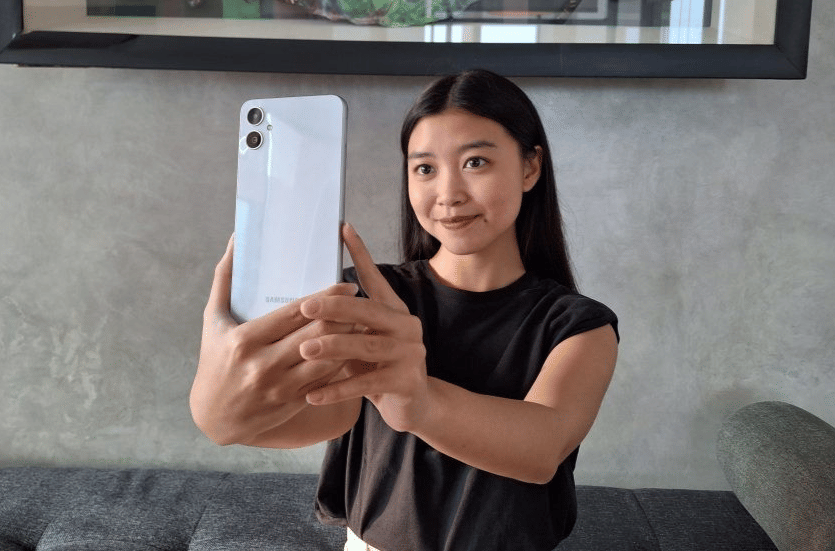 Seorang wanita memegang dan mengambil swafoto menggunakan ponsel Samsung Galaxy A05 berwarna perak di dalam ruangan.