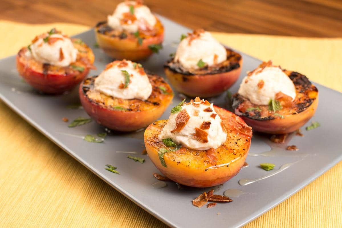 Beberapa potong peach panggang disajikan di atas piring abu-abu dengan topping krim dan taburan daun mint serta potongan karamel.