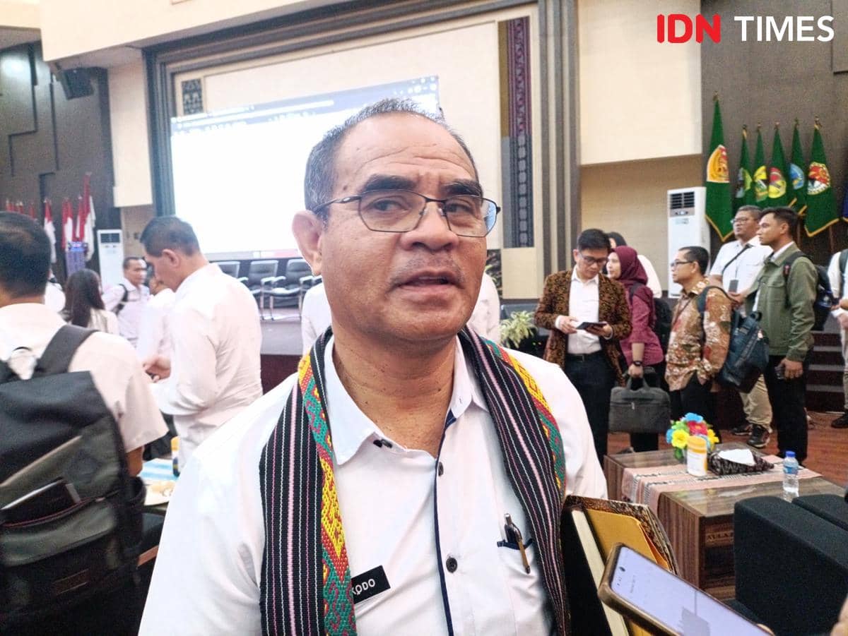 Seorang pria mengenakan kemeja putih dan selendang bermotif tenun sedang berada di dalam ruangan bersama sejumlah orang lain.