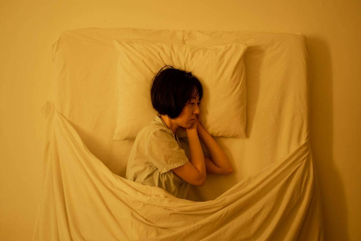 Seorang perempuan berbaring di tempat tidur dengan wajah tampak gelisah dan sulit tidur di bawah pencahayaan kuning redup.
