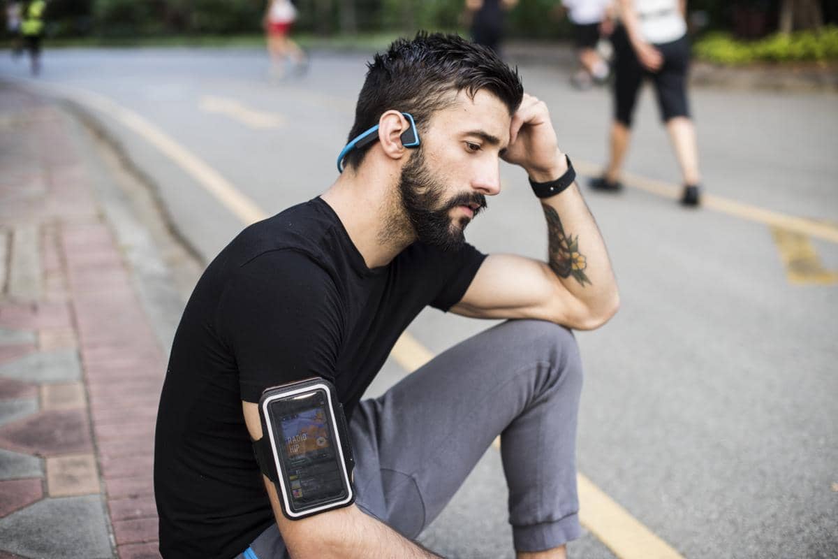 Seorang pelari duduk di trotoar sambil beristirahat, mengenakan earphone dan pelindung ponsel di lengan setelah berolahraga.