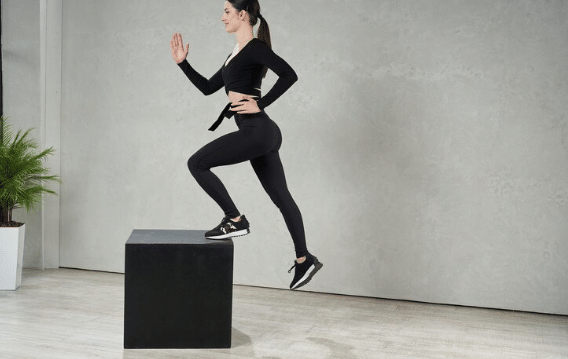 Seorang perempuan berolahraga melakukan latihan step-up di dalam ruangan dengan mengenakan pakaian olahraga hitam dan sepatu lari.