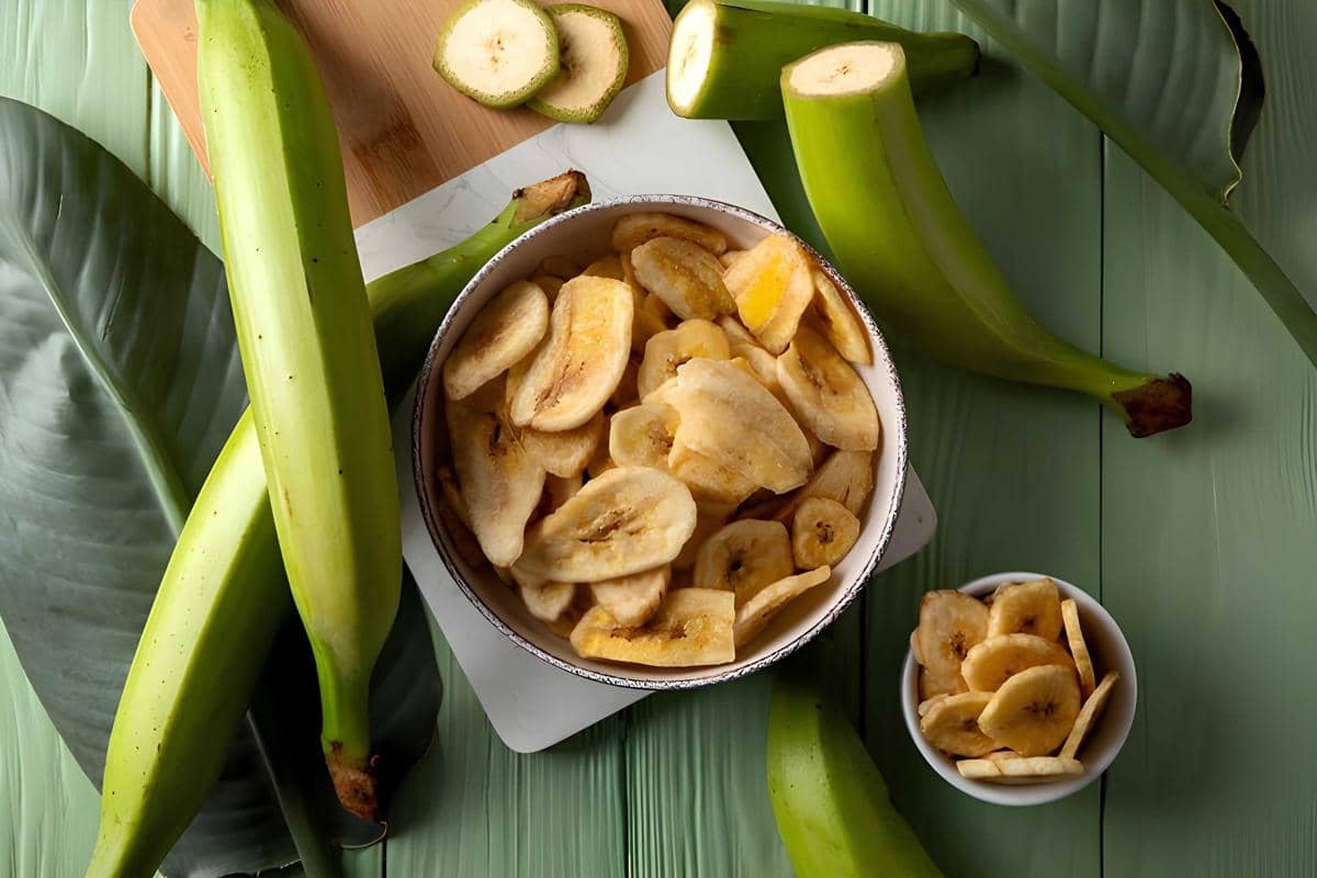 Keripik pisang renyah tersaji dalam mangkuk di atas meja hijau dengan pisang mentah dan daun pisang di sekitarnya.