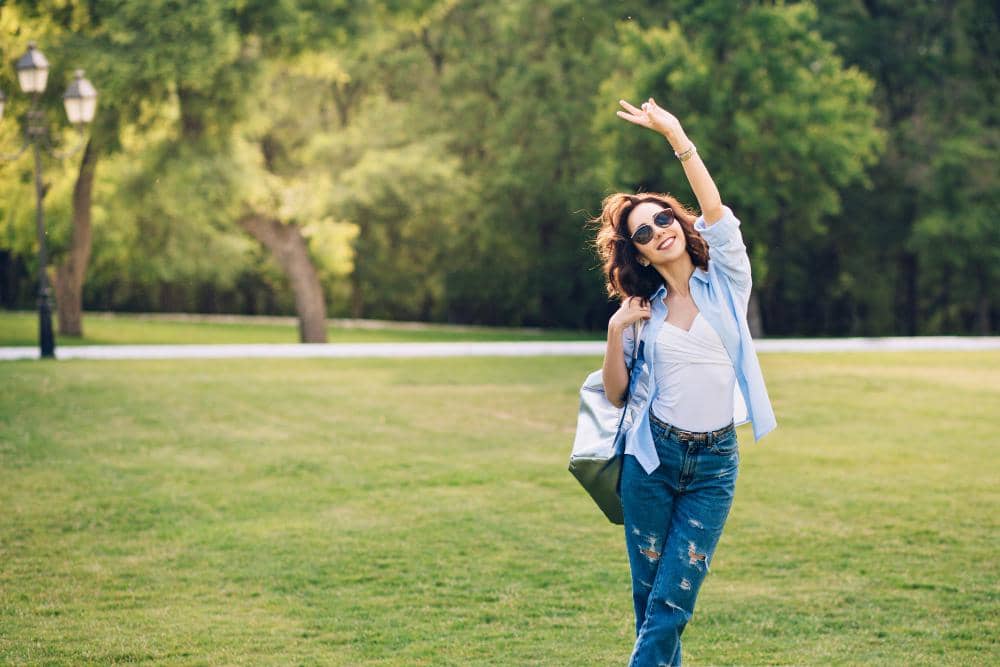 Perempuan mengenakan kemeja biru dan kacamata hitam tersenyum sambil mengangkat tangan di taman hijau yang cerah.