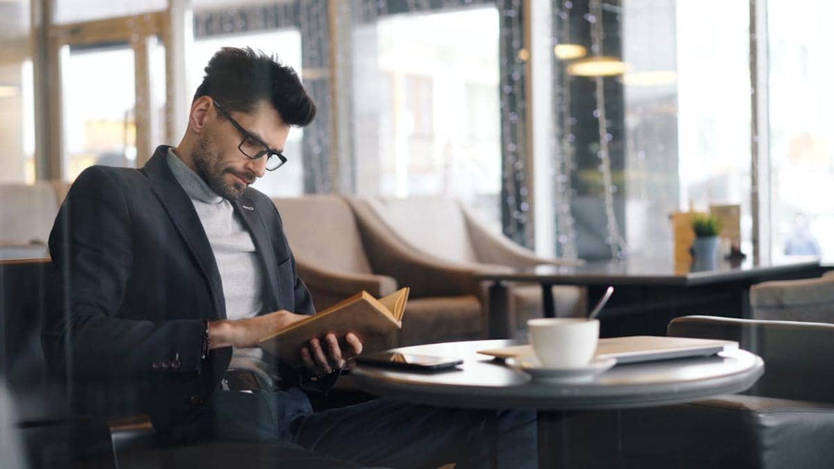 Seorang pria mengenakan jas dan kacamata sedang membaca buku di kafe dengan suasana tenang, di atas meja terdapat cangkir kopi dan tablet.