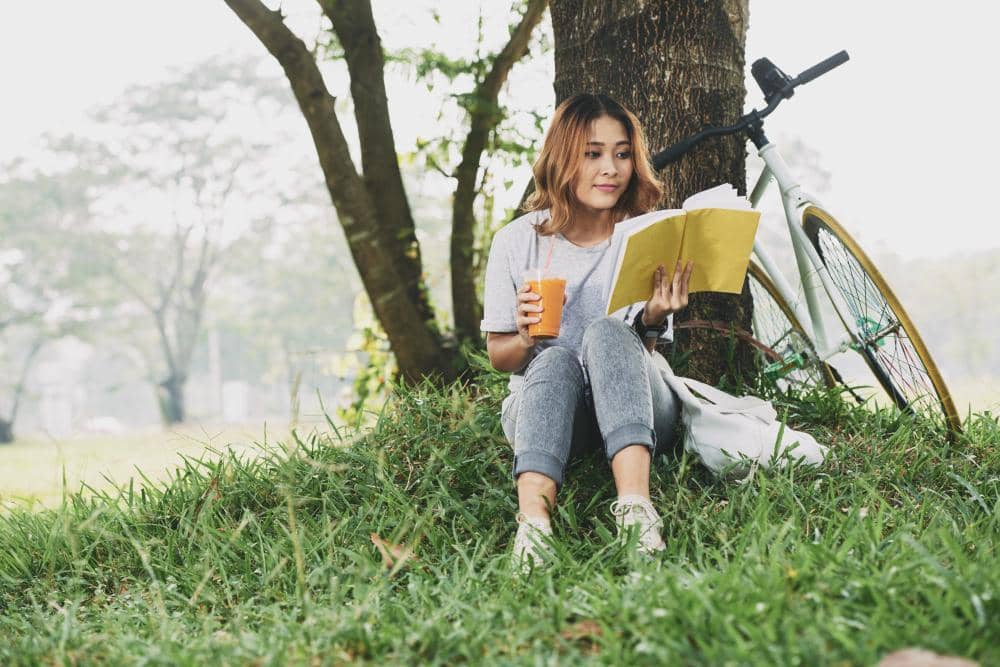 Seorang perempuan duduk bersandar di pohon sambil membaca buku dan memegang minuman di taman dengan sepeda di sampingnya.