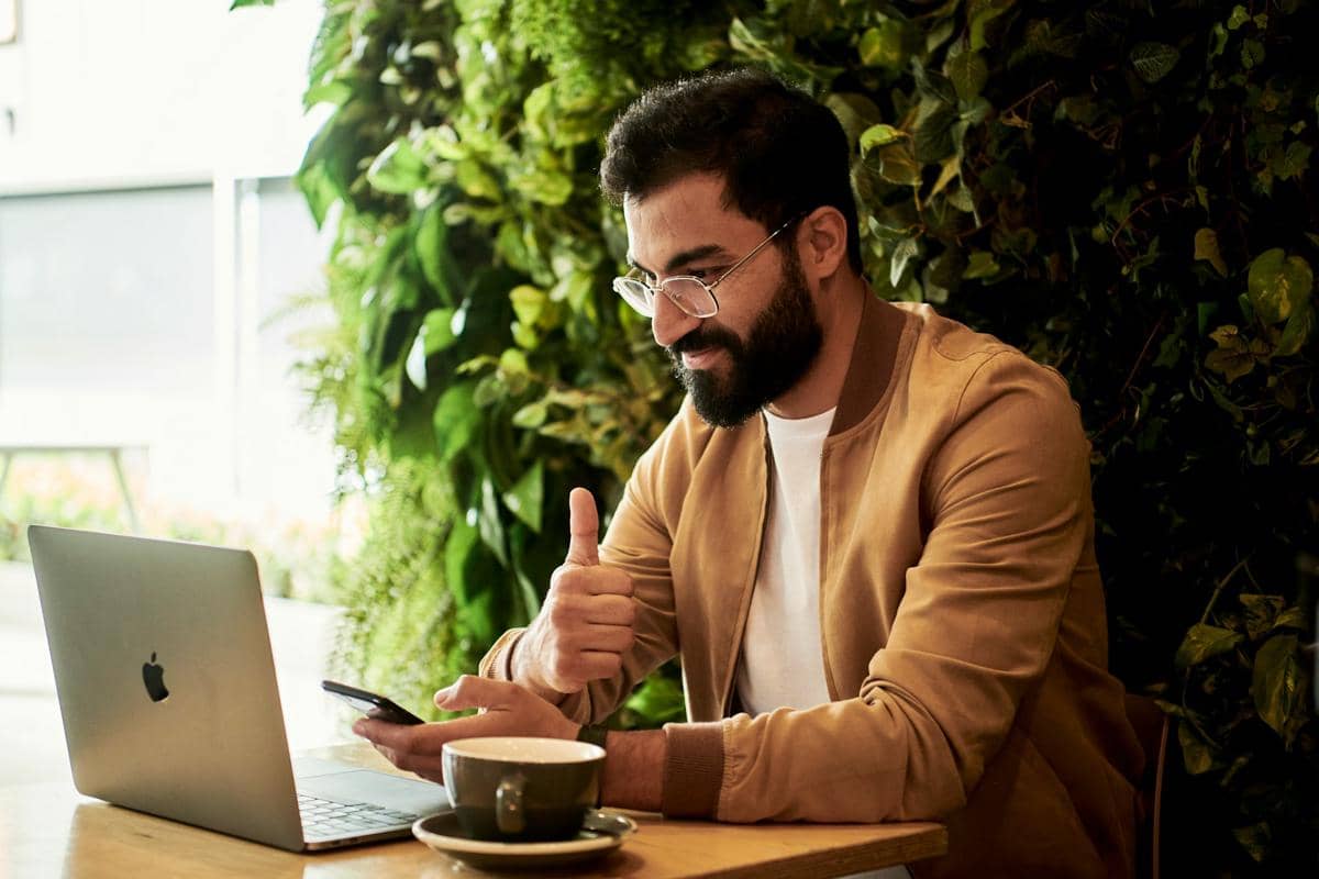 Seorang pria berjanggut mengenakan jaket cokelat duduk di kafe dengan laptop dan ponsel, mengacungkan jempol sambil tersenyum.