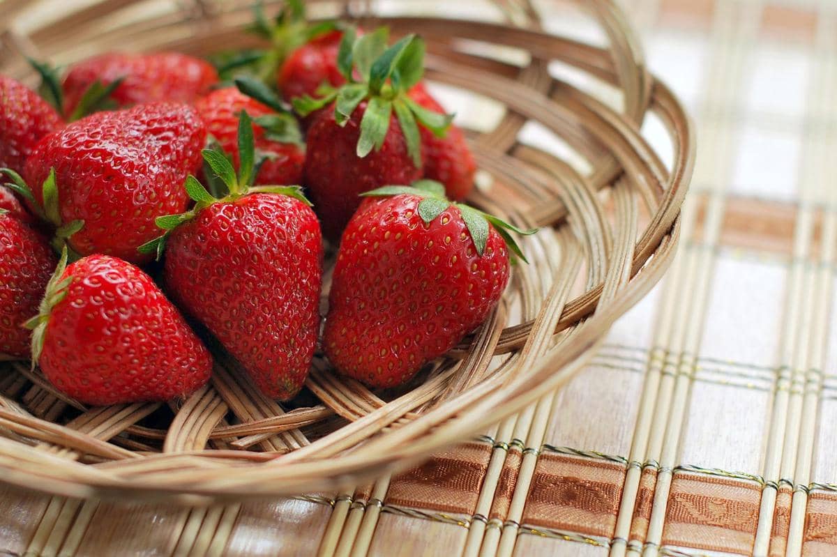 Beberapa stroberi segar berwarna merah cerah dengan daun hijau diletakkan di dalam keranjang anyaman di atas meja.