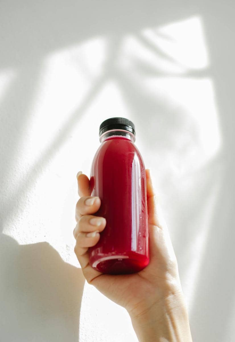 Tangan memegang botol minuman berwarna merah tua dengan tutup hitam di bawah cahaya alami di latar belakang putih.