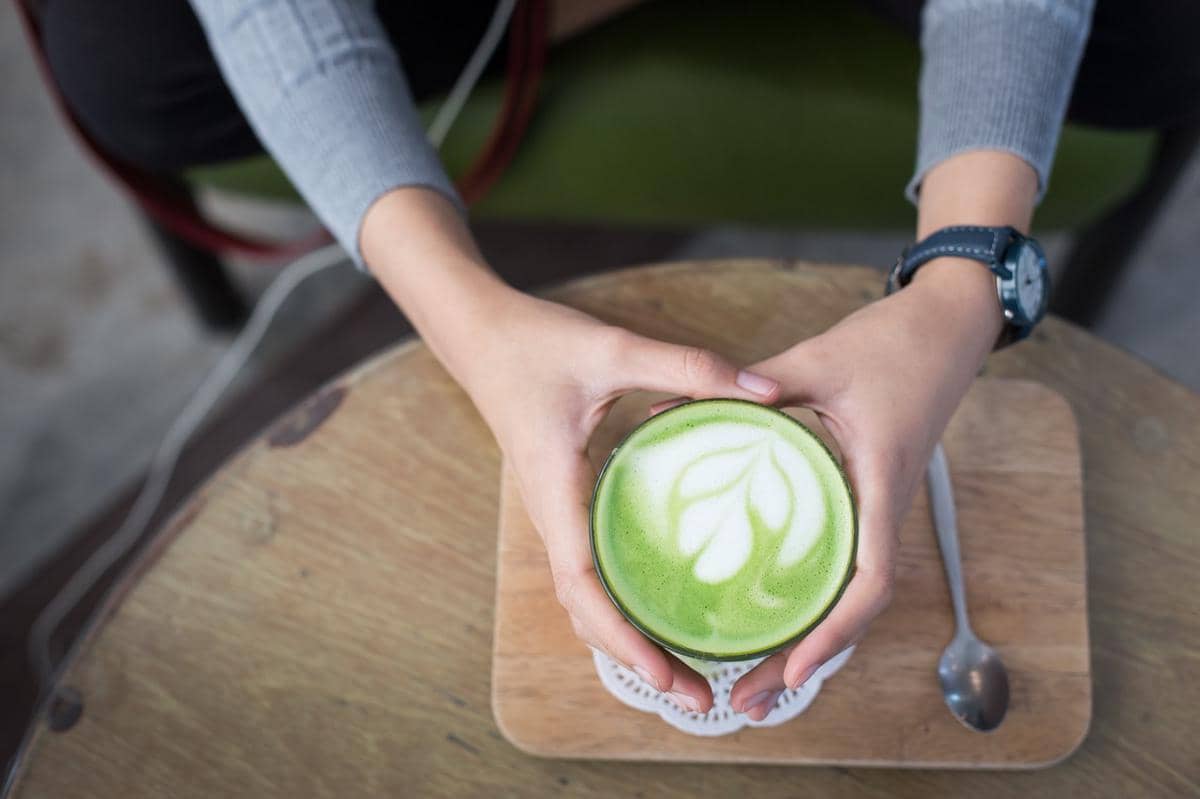 Ilustrasi seorang wanita berambut panjang menikmati minuman matcha sambil duduk santai di dalam ruangan.