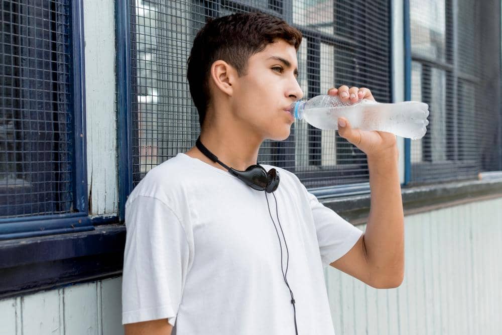 Seorang remaja mengenakan kaus putih dan headphone di leher sedang minum air dingin dari botol plastik di luar ruangan.