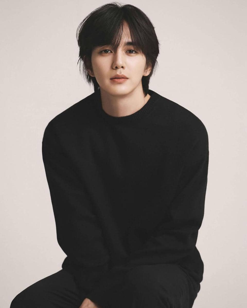 Seorang pria berambut hitam sedang duduk mengenakan sweter hitam dengan latar belakang polos berwarna terang.