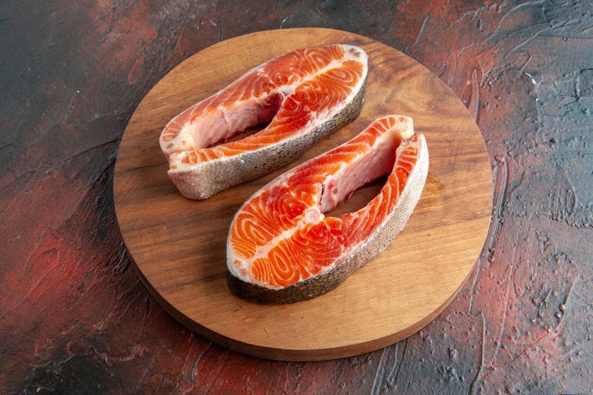 Dua potong daging ikan salmon mentah berwarna oranye dengan kulit perak di atas talenan kayu bundar.