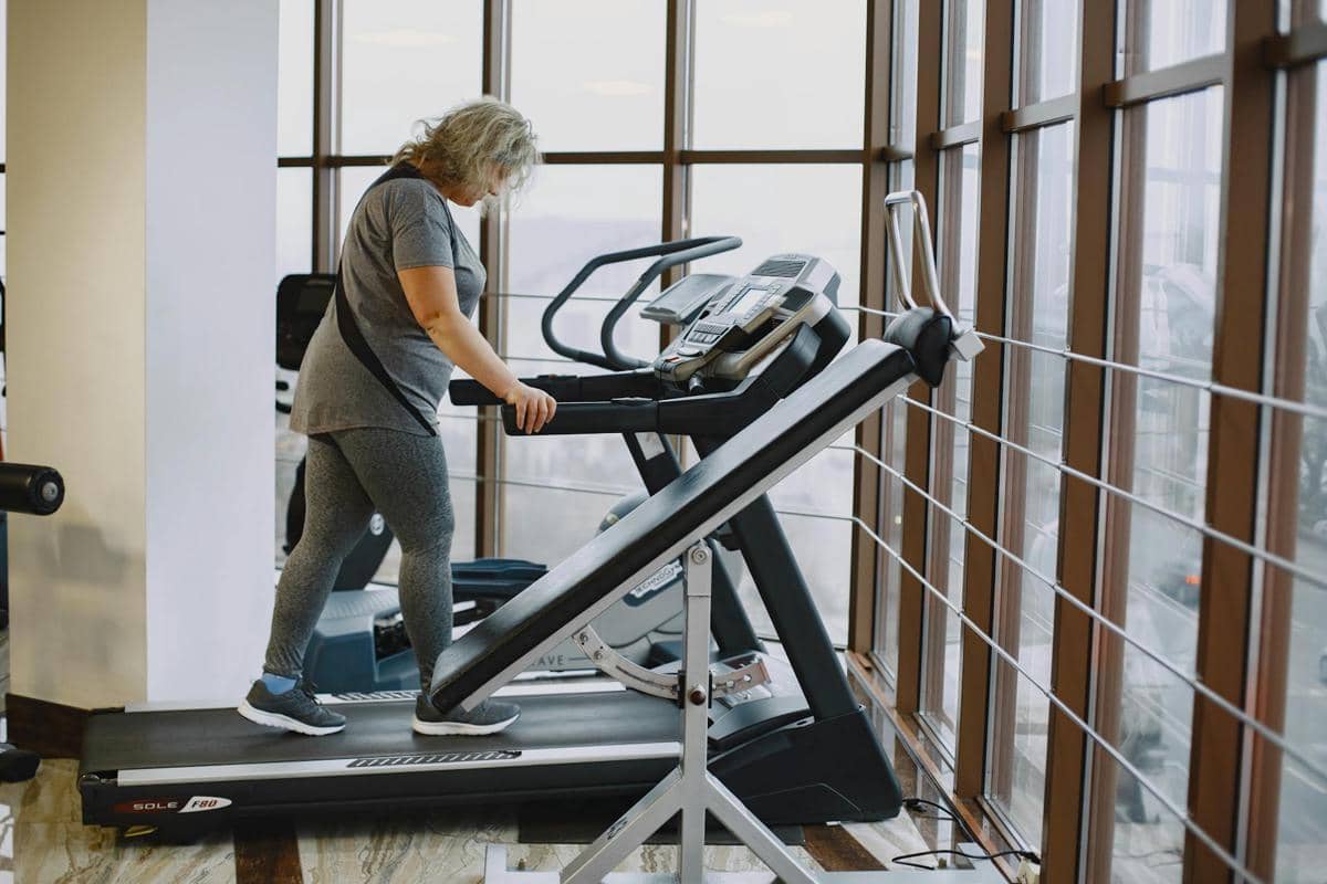 Seorang perempuan berjalan di atas treadmill dengan kemiringan di dalam ruangan, mengenakan pakaian olahraga.