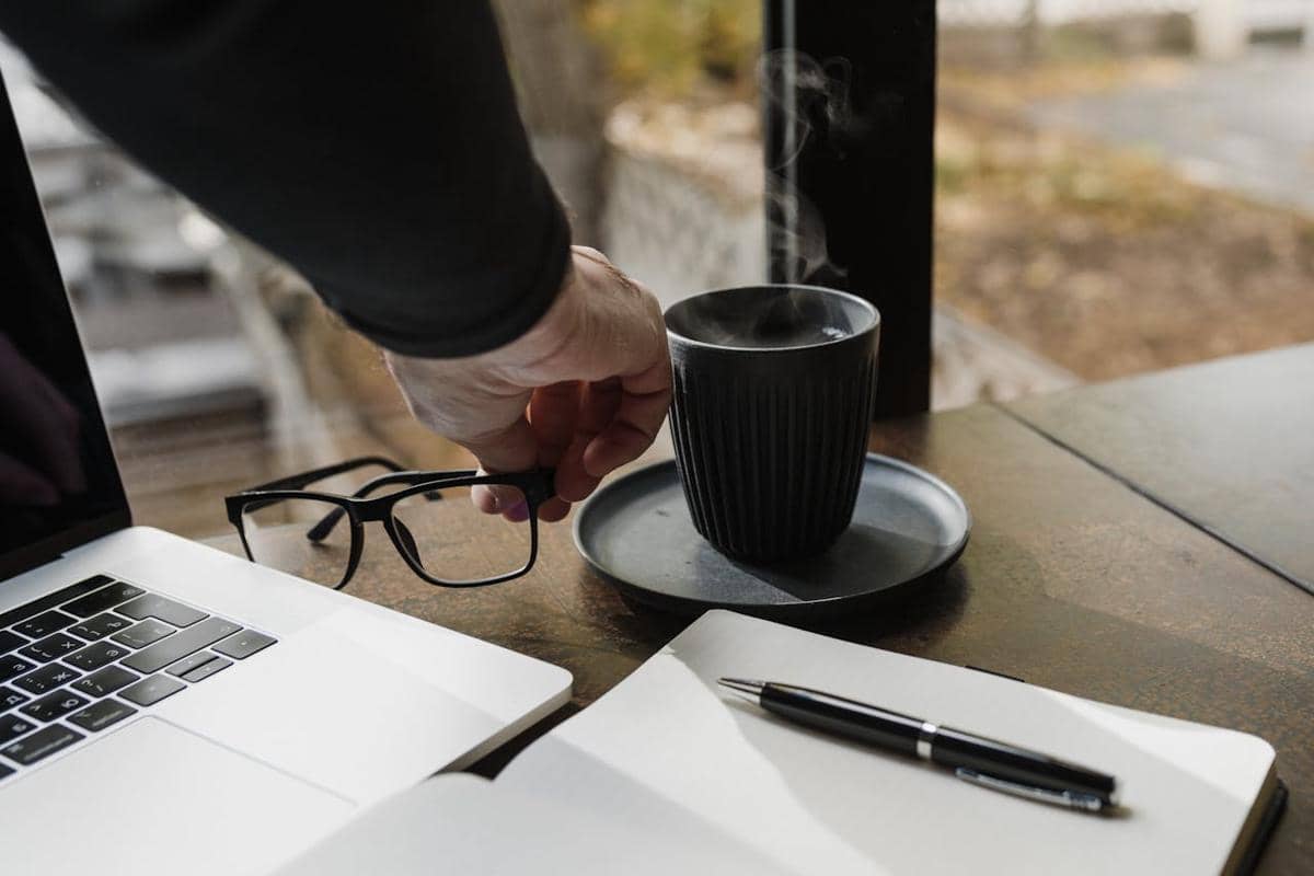 Tangan seseorang mengambil cangkir kopi di meja kerja dengan laptop, kacamata, buku catatan, dan pena di dekat jendela.