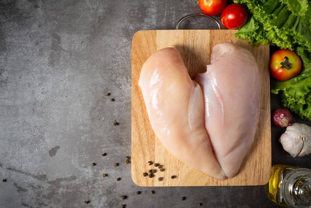 Dua potong dada ayam mentah di atas talenan kayu dengan tomat, selada, bawang putih, dan lada hitam di sekitarnya.