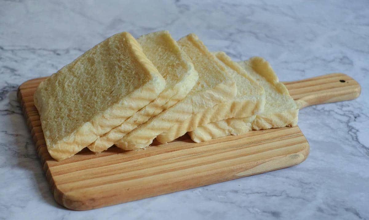 Beberapa potong roti tawar berwarna keemasan tersusun rapi di atas talenan kayu di permukaan meja marmer.