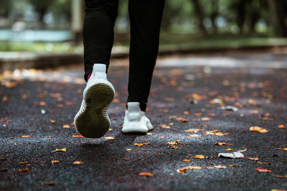Seseorang mengenakan sepatu lari putih berjalan di jalur taman beraspal yang dipenuhi daun kering saat suasana teduh.